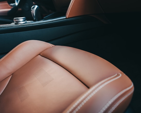 A close-up view of the interior of a luxury car, showcasing a brown leather seat with light stitching and perforated details. The background includes part of the car's dashboard and a metallic control knob, with accents of black and carbon fiber texture.