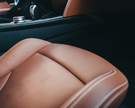 A close-up view of the interior of a luxury car, showcasing a brown leather seat with light stitching and perforated details. The background includes part of the car's dashboard and a metallic control knob, with accents of black and carbon fiber texture.