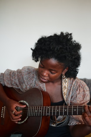 A person sitting and playing an acoustic guitar, deeply focused on the strings. The individual has dark curly hair and wears a patterned blouse. The background is neutral, drawing attention to the guitarist and the instrument.