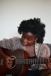 A person sitting and playing an acoustic guitar, deeply focused on the strings. The individual has dark curly hair and wears a patterned blouse. The background is neutral, drawing attention to the guitarist and the instrument.