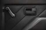 A close-up view of a car door interior featuring a black manual window crank and a door handle against a textured panel. The design includes geometric sections creating a modern aesthetic.