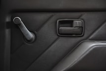 A close-up view of a car door interior featuring a black manual window crank and a door handle against a textured panel. The design includes geometric sections creating a modern aesthetic.