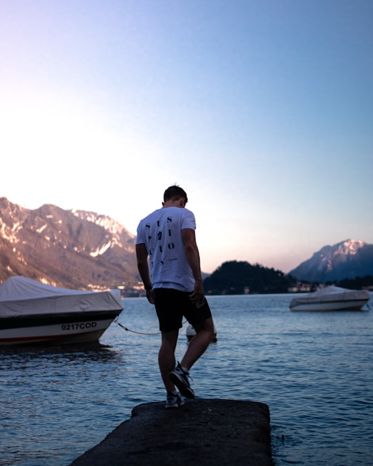 man in white shirt and black shorts standing on rock near body of water during daytime in Lake Como Italy