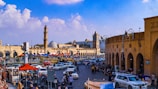 A bustling urban scene featuring a vibrant marketplace with numerous people, cars, and colorful vendor tents lining a busy street. A prominent ancient clock tower and a mosque with a large blue dome are visible in the background. Stalls and pedestrians fill the foreground, contributing to a lively atmosphere.