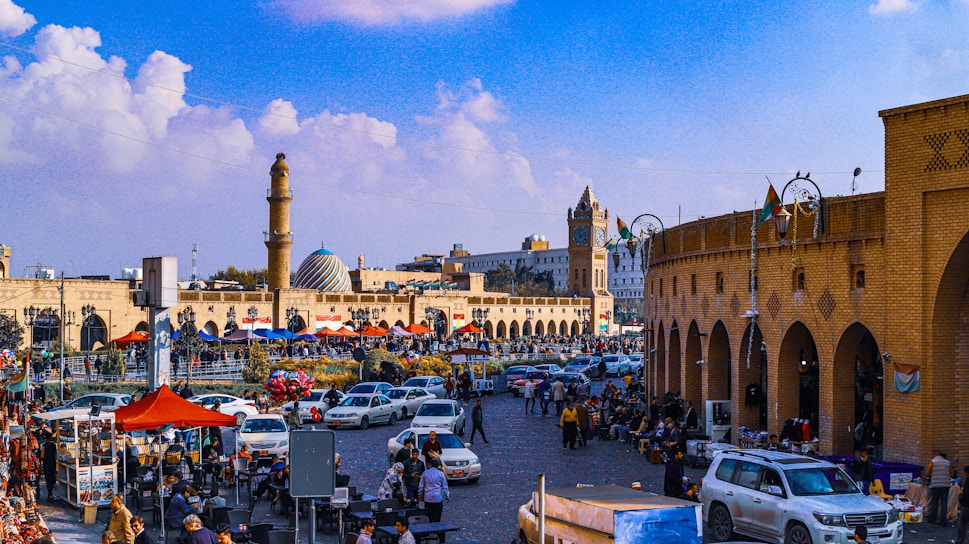 A vibrant street scene in an Arabic city bustling with people and colorful market stalls under a bright sky.