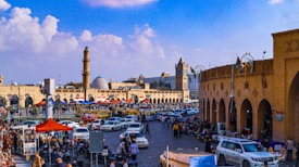 A bustling urban scene featuring a vibrant marketplace with numerous people, cars, and colorful vendor tents lining a busy street. A prominent ancient clock tower and a mosque with a large blue dome are visible in the background. Stalls and pedestrians fill the foreground, contributing to a lively atmosphere.