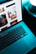 A laptop with a visible keyboard and screen displaying a webpage with various images, placed on a dark surface next to a glowing cylindrical light.
