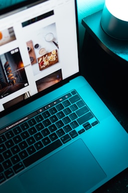 A laptop with a visible keyboard and screen displaying a webpage with various images, placed on a dark surface next to a glowing cylindrical light.