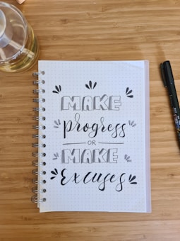 A wooden desk holds a spiral-bound notebook with dotted pages displaying calligraphy reading 'MAKE progress or MAKE excuses.' A transparent liquid-filled bottle and a black pen are placed nearby.