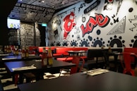 A trendy restaurant interior featuring a black and white theme with vibrant red accents. The walls are adorned with graffiti-style art including bold text, abstract shapes, and a large red character. Tables are set with ketchup bottles and utensils, while red and black chairs complement the decor. The lighting is warm and modern, enhancing the urban vibe. A television is mounted on one wall displaying a sports event.