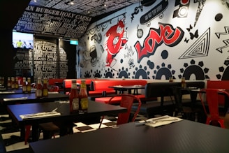 A trendy restaurant interior featuring a black and white theme with vibrant red accents. The walls are adorned with graffiti-style art including bold text, abstract shapes, and a large red character. Tables are set with ketchup bottles and utensils, while red and black chairs complement the decor. The lighting is warm and modern, enhancing the urban vibe. A television is mounted on one wall displaying a sports event.