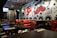 A trendy restaurant interior featuring a black and white theme with vibrant red accents. The walls are adorned with graffiti-style art including bold text, abstract shapes, and a large red character. Tables are set with ketchup bottles and utensils, while red and black chairs complement the decor. The lighting is warm and modern, enhancing the urban vibe. A television is mounted on one wall displaying a sports event.
