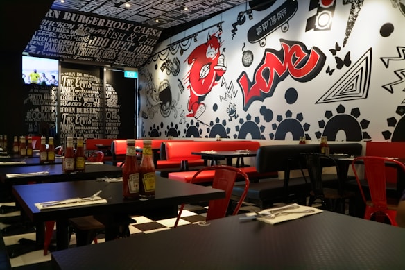 A trendy restaurant interior featuring a black and white theme with vibrant red accents. The walls are adorned with graffiti-style art including bold text, abstract shapes, and a large red character. Tables are set with ketchup bottles and utensils, while red and black chairs complement the decor. The lighting is warm and modern, enhancing the urban vibe. A television is mounted on one wall displaying a sports event.