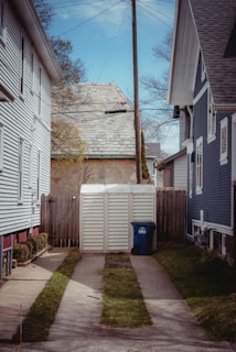 Between two houses with differing siding styles, a narrow path leads to a wooden shed surrounded by a wooden fence. A blue recycling bin is placed next to the shed. A telephone pole with wires stretches upwards, and the sky is partly cloudy. Trees without leaves are visible in the background.