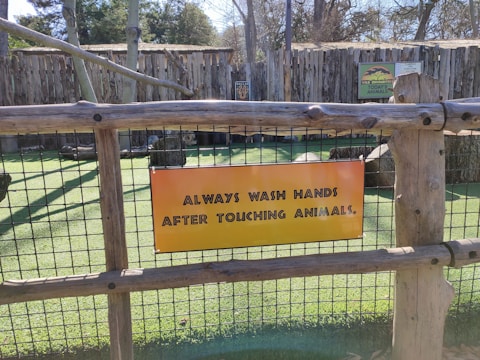 A wooden fence with wire mesh supports a bright yellow-orange sign which reads 'Always wash hands after touching animals.' Behind the fence, there are animal enclosures with green grass and wooden structures. There are additional signs on the structures in the background providing more information.