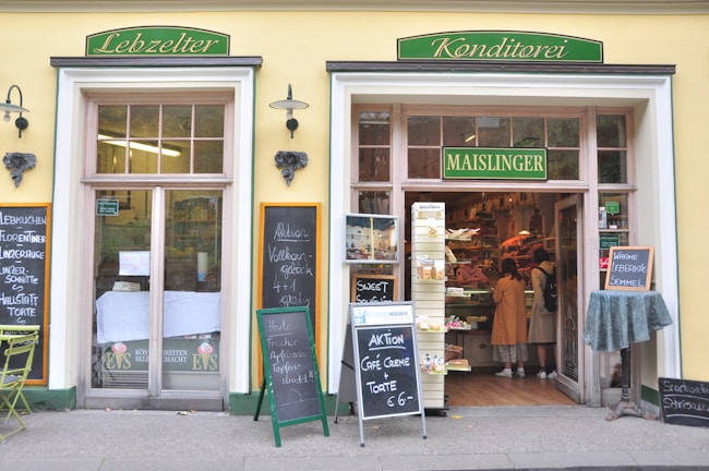 The image depicts the exterior of a traditional bakery and confectionery shop named 'Maislinger'. The storefront features two entrances with large windows displaying various signs and advertisements. The left entrance has a sign 'Lebzelter', while the right entrance is marked 'Konditorei'. There are blackboards with written menus and offers placed outside the shop. Two people are seen inside the shop, looking at the displayed baked goods. The shop exterior is painted yellow with green and gold signage.