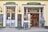 The image depicts the exterior of a traditional bakery and confectionery shop named 'Maislinger'. The storefront features two entrances with large windows displaying various signs and advertisements. The left entrance has a sign 'Lebzelter', while the right entrance is marked 'Konditorei'. There are blackboards with written menus and offers placed outside the shop. Two people are seen inside the shop, looking at the displayed baked goods. The shop exterior is painted yellow with green and gold signage.