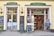 The image depicts the exterior of a traditional bakery and confectionery shop named 'Maislinger'. The storefront features two entrances with large windows displaying various signs and advertisements. The left entrance has a sign 'Lebzelter', while the right entrance is marked 'Konditorei'. There are blackboards with written menus and offers placed outside the shop. Two people are seen inside the shop, looking at the displayed baked goods. The shop exterior is painted yellow with green and gold signage.