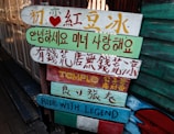 Colorful wooden signs with various languages, including Chinese, Korean, and English, are stacked on a pole. They are painted in bright colors with different messages and symbols.