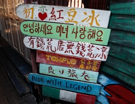 Colorful wooden signs with various languages, including Chinese, Korean, and English, are stacked on a pole. They are painted in bright colors with different messages and symbols.