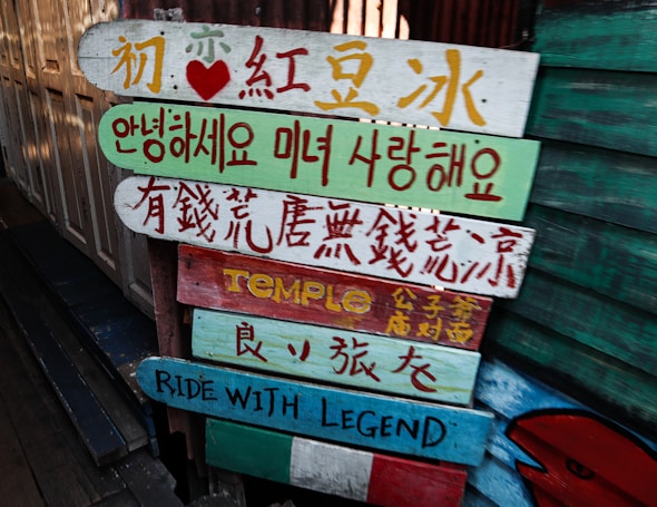 Colorful wooden signs with various languages, including Chinese, Korean, and English, are stacked on a pole. They are painted in bright colors with different messages and symbols.