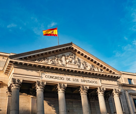 white concrete building with red and yellow flag on top under blue sky during daytime in Thyssen-Bornemisza Museum Spain