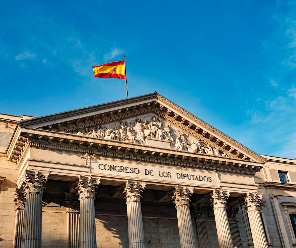 Edificio de hormigón blanco con bandera roja y amarilla en la parte superior bajo el cielo azul durante el día