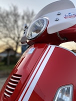 A close-up view of a red scooter, focusing on the front headlight and part of the windshield. The scooter features white racing stripes and a Vespa Servizio badge. The background shows a tree and a structure blurred out, indicating it was taken outdoors.