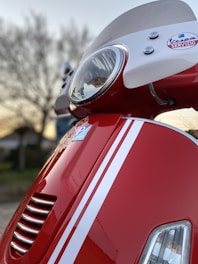A close-up view of a red scooter, focusing on the front headlight and part of the windshield. The scooter features white racing stripes and a Vespa Servizio badge. The background shows a tree and a structure blurred out, indicating it was taken outdoors.