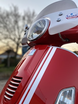 A close-up view of a red scooter, focusing on the front headlight and part of the windshield. The scooter features white racing stripes and a Vespa Servizio badge. The background shows a tree and a structure blurred out, indicating it was taken outdoors.