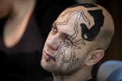 A person with artistic, intricate black designs painted on their face and scalp is seen looking slightly upward. The designs consist of abstract patterns and shapes that cover a significant portion of their head.