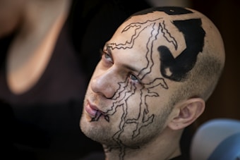 A person with artistic, intricate black designs painted on their face and scalp is seen looking slightly upward. The designs consist of abstract patterns and shapes that cover a significant portion of their head.