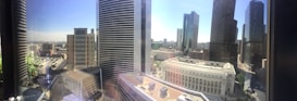 A panoramic view of a cityscape filled with tall skyscrapers under a clear blue sky. The foreground features a modern office building with reflective windows, while the background consists of several high-rise buildings and a sprawling urban area. The sunlit scene includes visible roads and traffic, surrounded by architectural structures of varying designs.