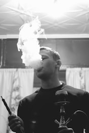 A person is smoking a hookah, with a large plume of smoke rising in the air. The setting appears indoors, with curtains and a lit ceiling fixture visible in the background. The image is in black and white, creating a classic or vintage feel.