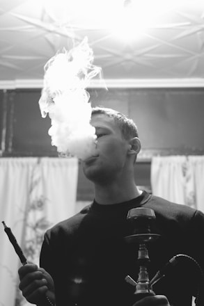 A person is smoking a hookah, with a large plume of smoke rising in the air. The setting appears indoors, with curtains and a lit ceiling fixture visible in the background. The image is in black and white, creating a classic or vintage feel.