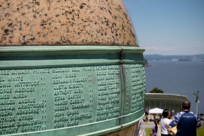 A detailed, close-up view of a war memorial with engraved names on a smooth, greenish metal surface and a rounded stone top. In the background, people are visible outdoors, facing a scenic view of a large body of water and distant hills under a clear blue sky.