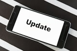 A smartphone displaying the word 'Update' on its screen is placed on a black and white striped surface. The phone is in landscape orientation, and the image focuses on the screen’s content.