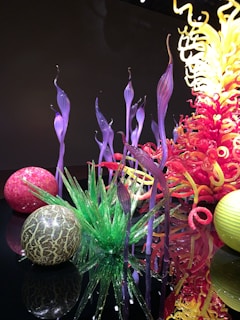 A vibrant and intricate glass sculpture composed of swirling purple, red, and yellow forms, along with spherical glass elements. The pieces are artistically arranged, creating a visually dynamic display with reflections on a glossy black surface.