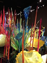An intricate display of colorful glass sculptures and structures with elongated forms and vibrant patterns. The art pieces include tall, thin spires in red and orange, as well as broad, fan-like shapes in blue and green. Yellow bowl-shaped forms add contrast to the scene.