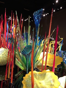 An intricate display of colorful glass sculptures and structures with elongated forms and vibrant patterns. The art pieces include tall, thin spires in red and orange, as well as broad, fan-like shapes in blue and green. Yellow bowl-shaped forms add contrast to the scene.