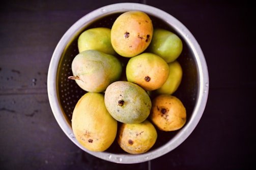 A metal colander containing a variety of ripe and unripe mangoes sits against a dark background. The mangoes display different hues of yellow, orange, and green, with some showing brown spots and a textured surface.