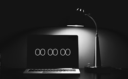 A minimalist setup with a laptop displaying a digital clock set at '00:00', beside a modern desk lamp casting a soft glow over the scene, creating contrast in an otherwise dark environment.