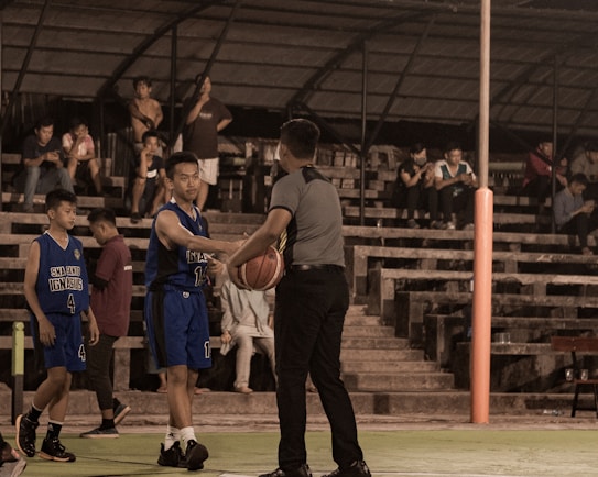 Several people are gathered at a basketball court, with a focus on a referee handing a basketball to a player wearing a blue jersey. Spectators are seated on the steps in the background, some watching intently while others are on their phones. The setting appears to be a dimly lit indoor or outdoor court.