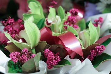 A vibrant floral arrangement featuring pale green orchids and rich pink flowers, accented with broad green leaves and red ribbon. The composition creates a lush and colorful display with delicate textures and colors.