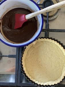 A pot containing melted chocolate with a red spatula inside is placed on a stove. Next to it, there is an unbaked tart crust in a fluted tart pan.