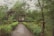 A rustic house with a thatched roof is nestled amidst dense greenery. A gravel path lined with vibrant red flowers and lush shrubs leads up to the elevated structure. Large trees surround the area, providing a serene and natural setting.