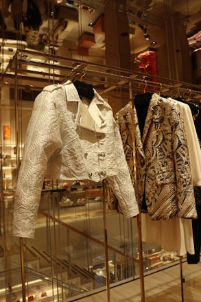 A close-up view of a fashion display featuring intricately designed jackets hanging on a rack in an upscale retail environment. The jackets exhibit detailed patterns and textures, with one appearing in a white hue with embossed geometric designs, and the other showcasing a gold and black intricate pattern. The background shows a reflection of the luxurious store interior, with shelves, lighting, and other merchandise visible.