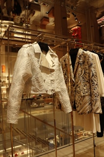 A close-up view of a fashion display featuring intricately designed jackets hanging on a rack in an upscale retail environment. The jackets exhibit detailed patterns and textures, with one appearing in a white hue with embossed geometric designs, and the other showcasing a gold and black intricate pattern. The background shows a reflection of the luxurious store interior, with shelves, lighting, and other merchandise visible.