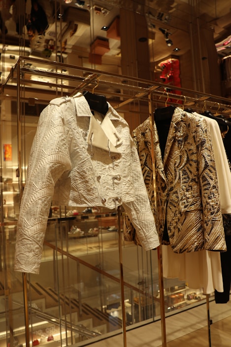 A close-up view of a fashion display featuring intricately designed jackets hanging on a rack in an upscale retail environment. The jackets exhibit detailed patterns and textures, with one appearing in a white hue with embossed geometric designs, and the other showcasing a gold and black intricate pattern. The background shows a reflection of the luxurious store interior, with shelves, lighting, and other merchandise visible.