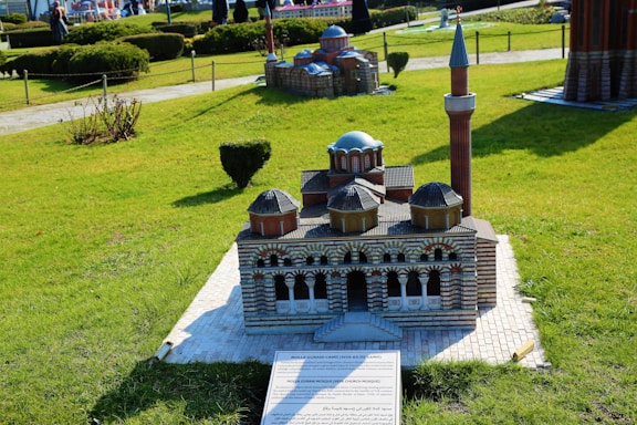 A detailed model of a mosque with distinctive domes and a minaret, set against a grassy landscape with hedges and pathways. The model shows intricate architectural features, including a series of arches in the facade and a mix of brick colors. A plaque with text is positioned in front of the model.